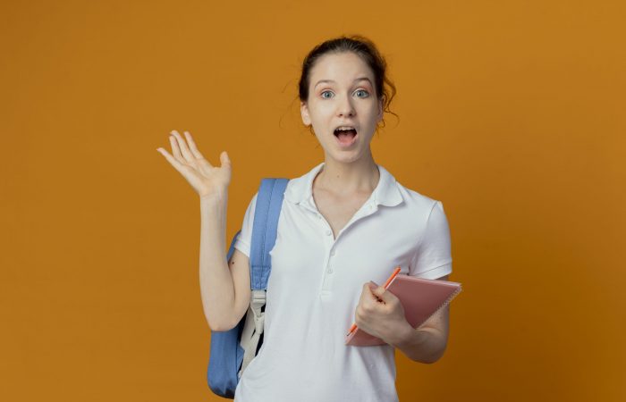 impressed young pretty female student wearing back bag holding note pad and pen showing empty hand isolated on orange background with copy space